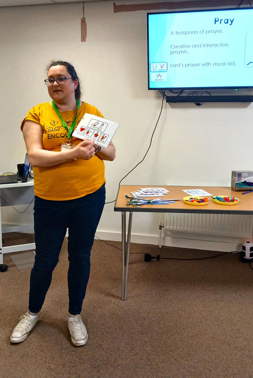 Female wearing jeans and a yellow tshirt, standing talking with a screen that says Pray behind her