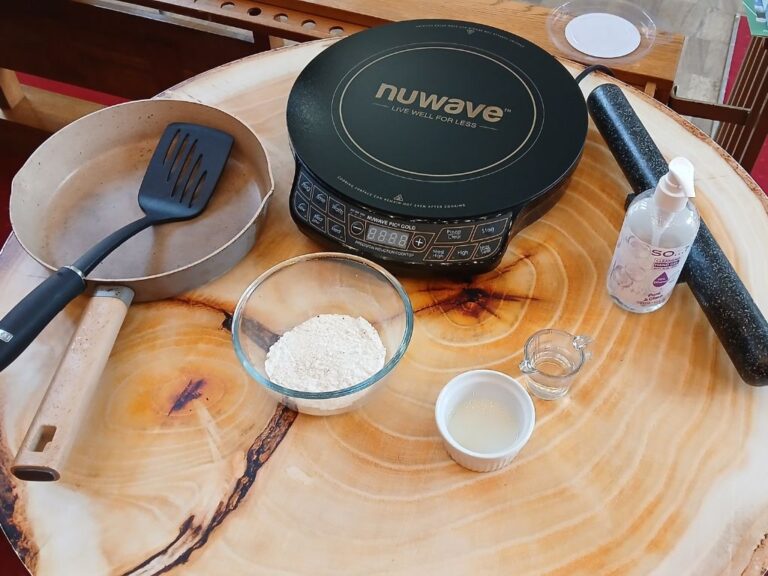 A small induction hob, frying pan, spatula, small bowl of flour, oil and water on a table.