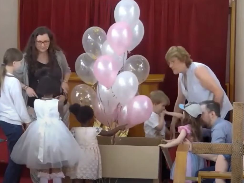 A group of people are trying unsuccessfully to return a large bunch of helium balloons to a box.