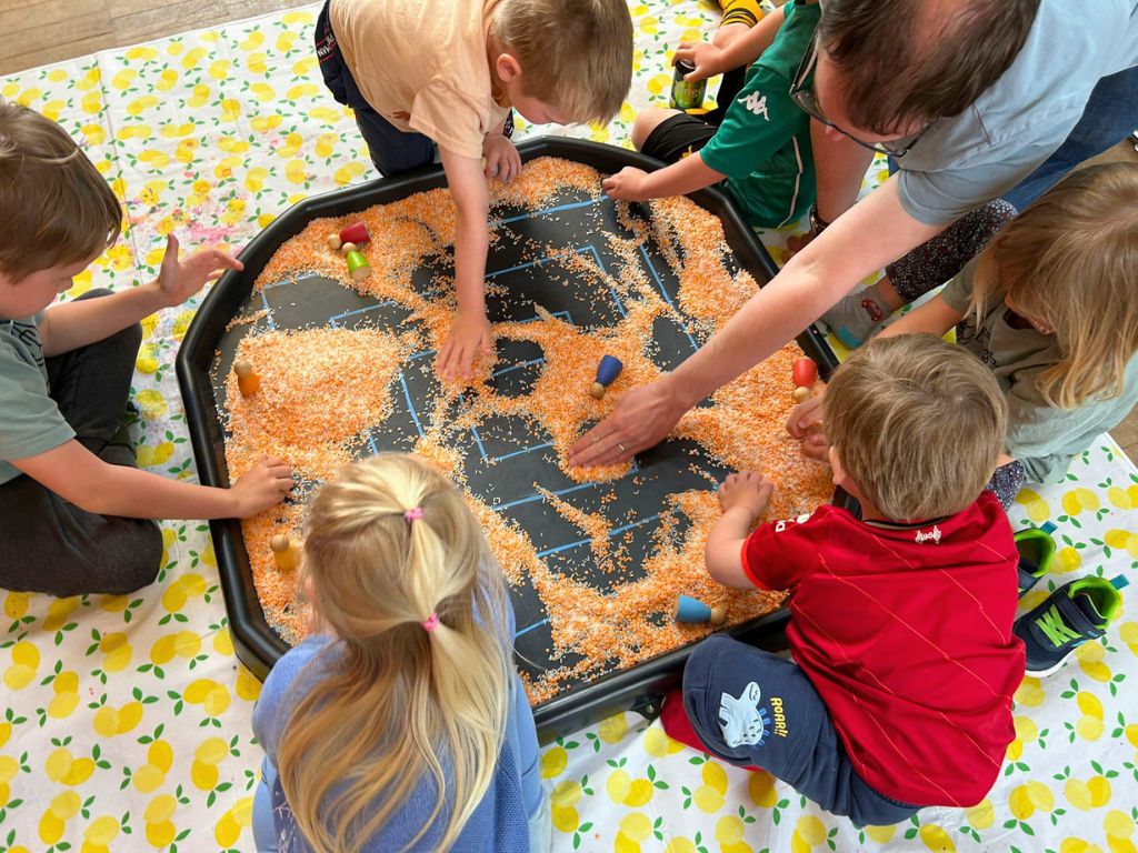 A tuff tray is on the ground. On the tuff tray is a drawn labyrinth partially covered by lentils. A group of childen and adults are uncovering the route and using peg dolls to travel around it.