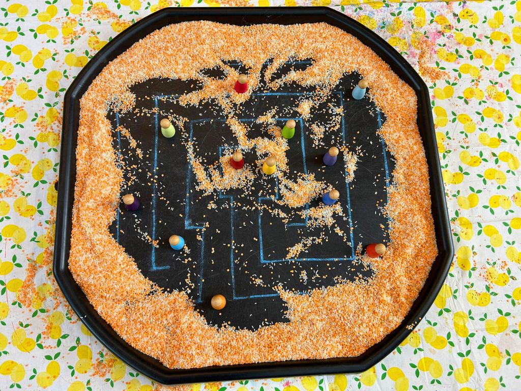 A tuff tray is on the ground. On the tuff tray is a drawn labyrinth partially covered by lentils. Several peg dolls are arranged on the path of the labyrinth.