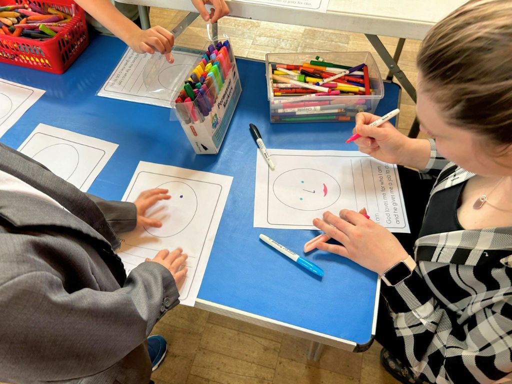 Adults and children are drawing their faces on pieces of paper.