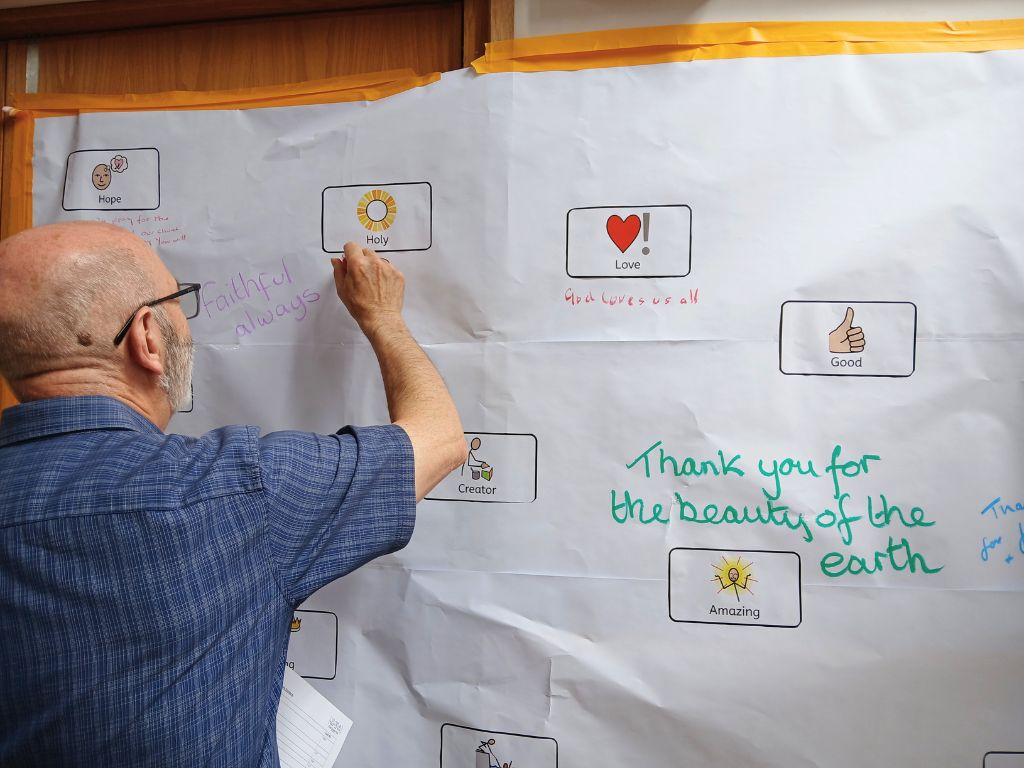A man is writing on a wall covered in white paper. On the paper are words of adoration that describe God and people have written their own prayers.