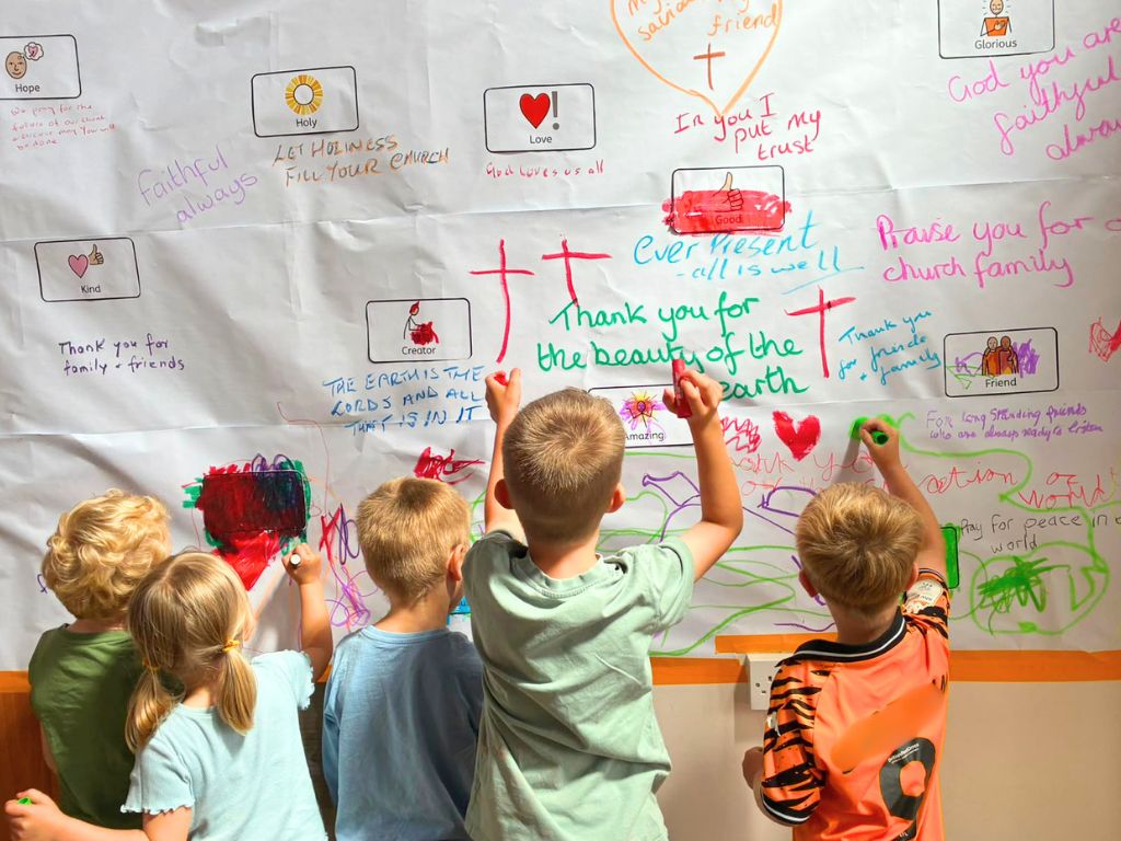 A group of children adding their prayers to a paper graffiti wall. The wall is covered in prayers people have written in coloured pens. These include 