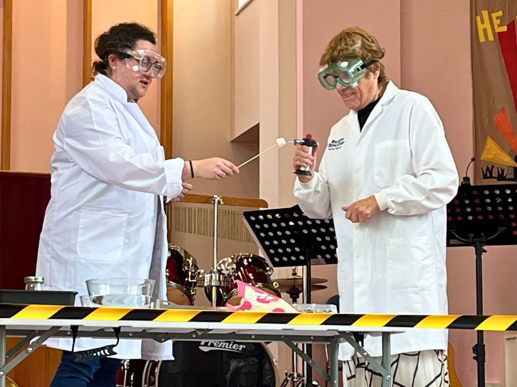 Two women in lab coats and safety goggles are stood behind a hazard tape barrier. One is holding a chef's blowtorch and the other is toasting a marshmallow on a skewer.