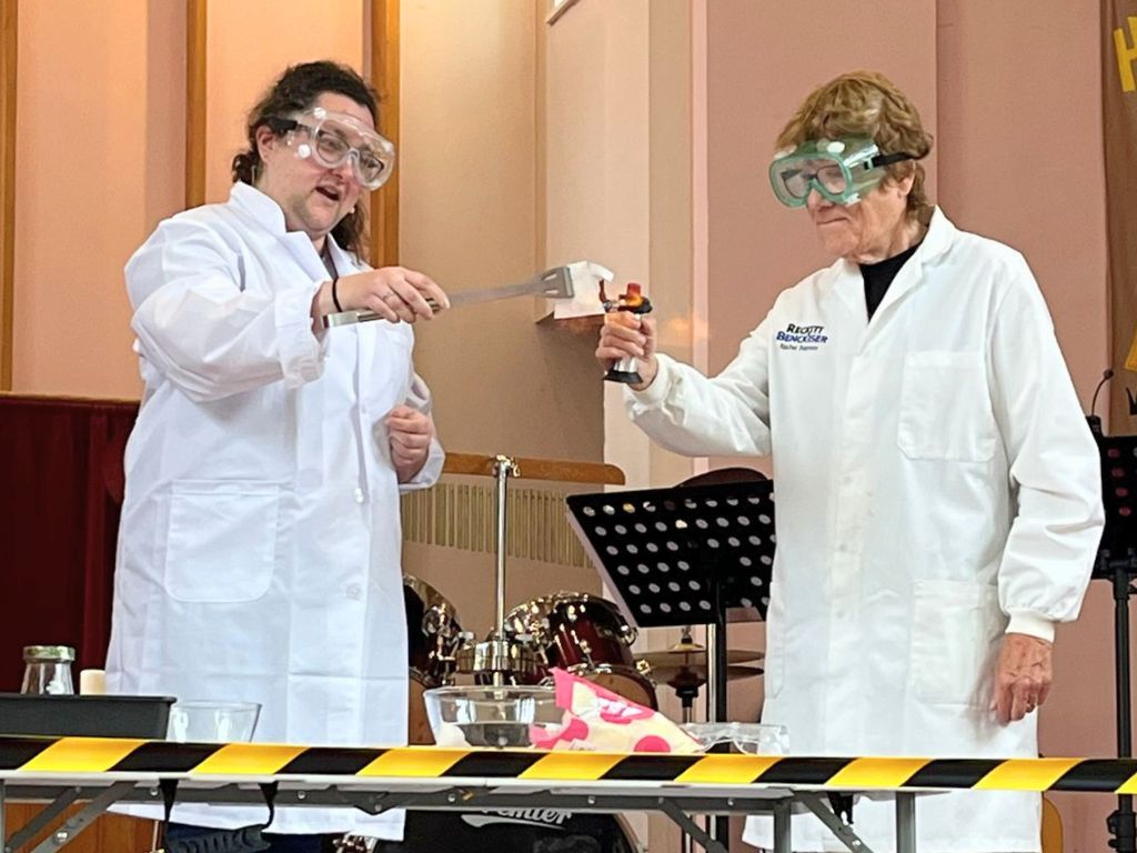 Two women in lab coats and safety goggles are stood behind a hazard tape barrier. One is holding a chef's blowtorch and the other is holding a piece of paper. Flames are around the paper but it is not singed.
