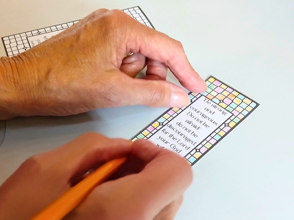 An adults hands are seen colouring in a bookmark with the words of Joshua 1:9.