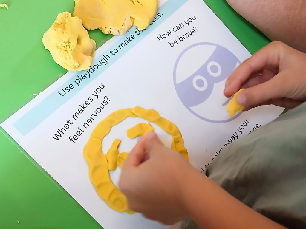 A child's hands are placing yellow play dough onto an outline of nervous and brave faces. On the mat are the questions 