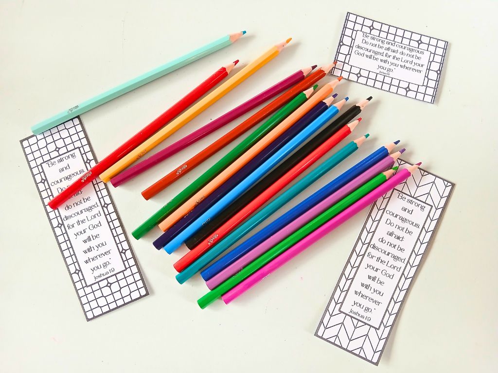 A selection of colouring pencils on a table. Around them are some bookmarks and wallet-sized cars with Joshua 1:9 written on them.