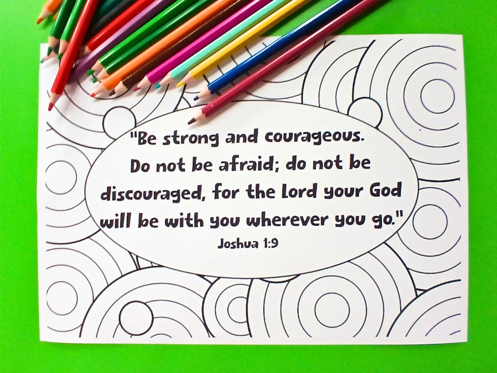 A large colouring sheet on a green table. The sheet contains the Bible verse Joshua 1:9. There are some coloured pencils on the table too.